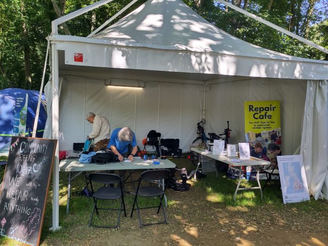 A photo of a gazeebo with tables inside and signs for a repair cafe.