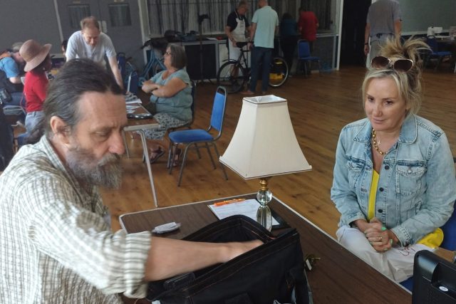 A photo of two people sitting at a table on which stands a lamp. One person is reaching into a toolkit.